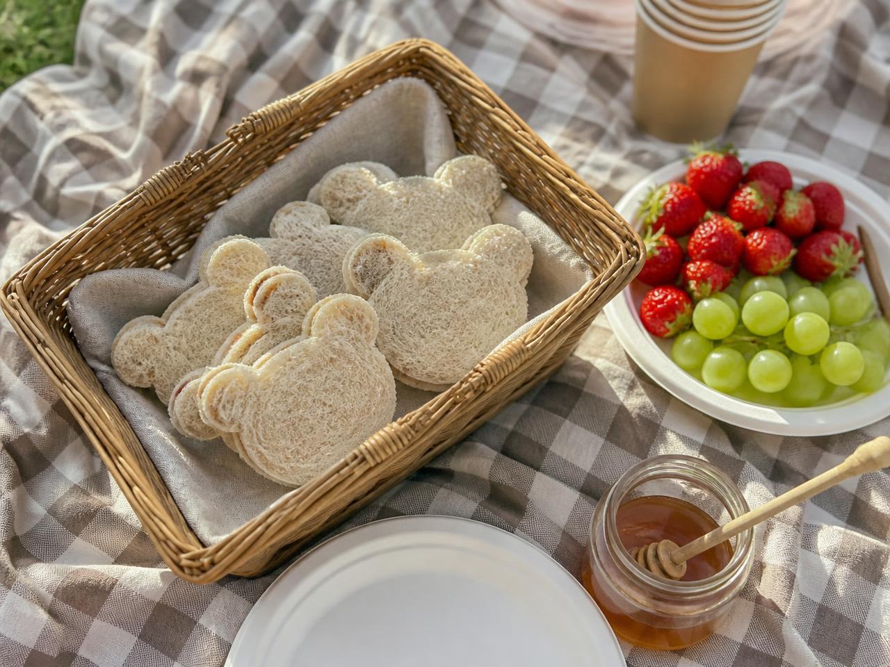 Bear-cutter sandwiches in a square picnic basket, with strawberries, grapes and honey.